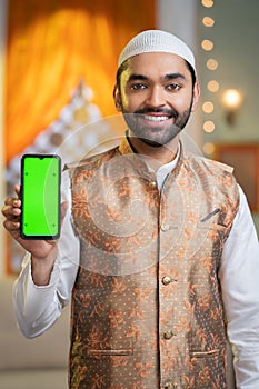 Vertical shot of happy young muslim man showing green screened mobile phone by looking at camera at home