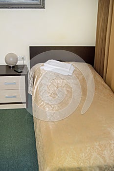 Vertical shot of the interior of the hotel bedroom with an empty single bed with wooden headboard and bedside table and towels on