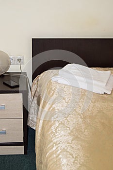 Vertical shot of the interior of the hotel bedroom with an empty single bed with wooden headboard and bedside table and towels on