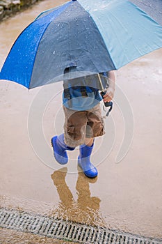 Vertical shot of a little boy with an umbrella and blue rubber boots walking through the wet ground