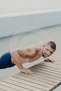 Vertical shot of muscular bearded man stands in plank pose trains chest and arms muscles has hard workout for keeping athletic