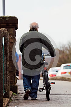 Vertical shot of an old man carrying a bicycle with him while walking on the sidewalk