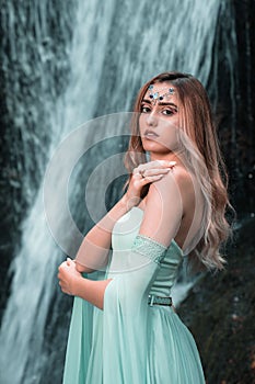 Vertical shot of the pretty woman wearing a long Tiffani dress and standing near the waterfall