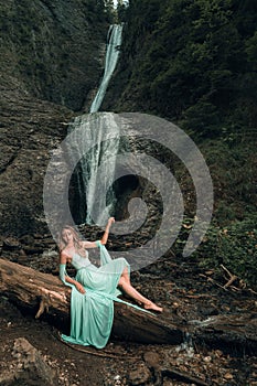 Vertical shot of the pretty woman wearing a long Tiffani dress and standing near the waterfall