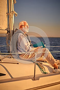 Vertical shot of relaxed retired man reading book and enjoying amazing sunset while sitting on the side of his sailboat