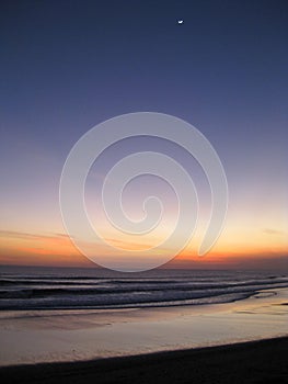 Vertical shot of the sea under a sky with a half moon during sunset