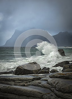 Vertical shot of the sea waves hitting the rocks with a mountain in clouds in the background