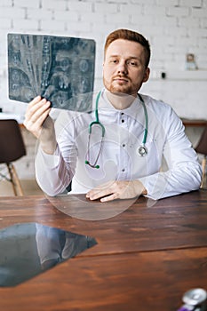 Vertical shot of serious male doctor in white uniform examining brain computerized tomography scan sitting at desk in