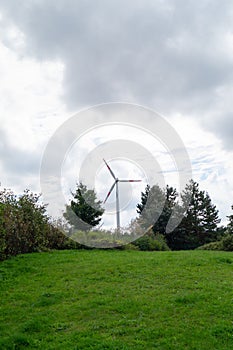 Vertical shot of a single wind turbine surrounded by trees and greenery against the cloudy sky