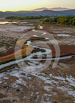 Vertical shot of a small twisty river flowing near a forest and green hills at sunset