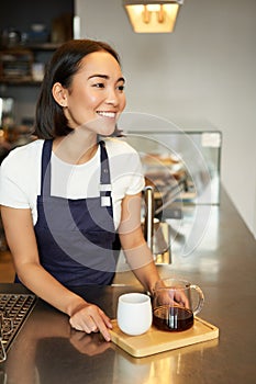 Vertical shot of smiling girl barista serving coffee, making batch brew, filter for client in cafe, wearing blue apron