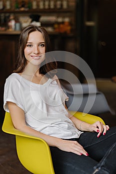 Vertical shot of smiling woman has dark hair, toothy smile, dressed in white t shirt, sits in yellow armchair, has red manicure, f