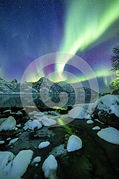 Vertical shot of the snow covered stones by a lake under the beautiful colorful northern lights