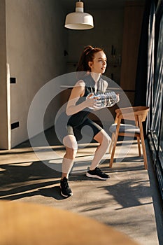 Vertical shot of sporty woman doing squat exercise holding in hands bottle with water. Athletic fit female wearing
