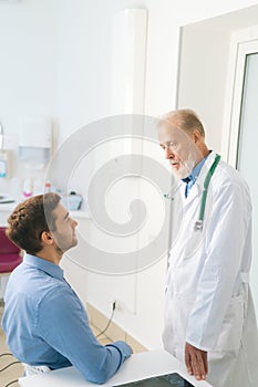 Vertical shot of standing mature male adult doctor examining male patient sitting at table in medical office.