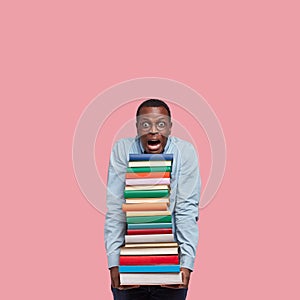 Vertical shot of stupefied black man leans head on pile of books, wears round glasses, surprised with many tasks for