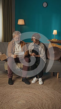 Vertical shot of a teenager, young man sitting with his grandfather on a sofa, couch, reading a document, paper