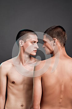 Vertical shot of two young caucasian half naked twin brothers looking at each other while standing isolated over grey