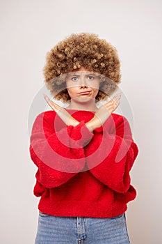 Vertical shot of upset young woman with curly hair keeps arms crossed with sad expression shows stop gesture, wears red