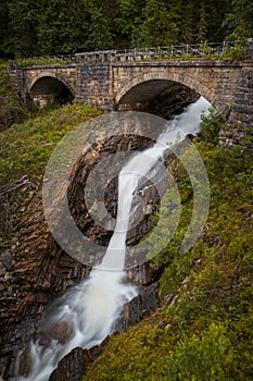 Vertical shot of a waterfall under bridge surrounded by greenery and rocks