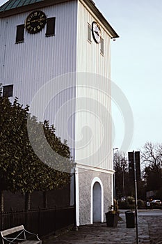 Vertical shot of a white building with windows and black clock, golden numbers, trees behind fence