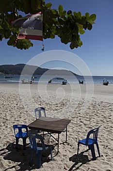 Vertical shot of a wooden table, four chairs standing in yellow sand under a green tree branch and a flag of Thailand at a Phuket