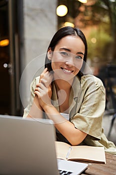 Vertical shot of young Asian woman working on remote from outdoor cafÃÂ©, sitting with laptop and smiling, studying