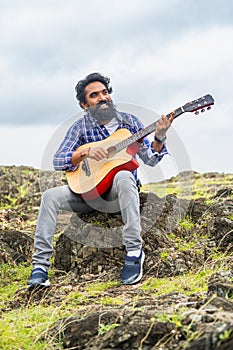 vertical shot of Young beard man playing guitar by singing song while sitting on top of hill - concept of hobbies