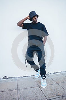 Vertical shot of a young black man in street-style clothes posing leaning on a white wall outdoors