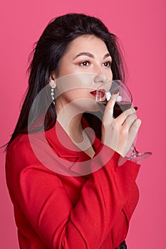 Vertical shot of young celebrating beautiful Caucasian woman wearing red elegant dress holding wine glass, enjoying its taste,