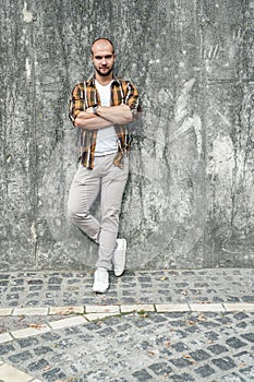 Vertical shot of young good looking bold bearded guy standing outdoors against grey modern loft wall with his arms crossed and