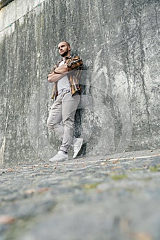 Vertical shot of young good looking bold bearded guy standing outdoors against grey modern loft wall with his arms crossed and