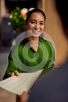 Vertical shot of a young happy asian female office worker giving some documents to her colleague and smiling while