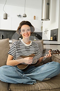 Vertical shot of young musician, woman learns how to play ukulele, sits on sofa with crossed legs and smiling at camera