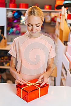 Vertical shot of young sales woman holding and using golden ribbon to tie bow for red wrapped gift box for Christmas