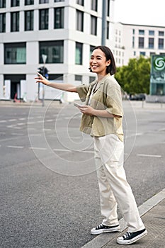 Vertical shot of young woman trying catch taxi, waving at driver on road, holding smartphone with car sharing app