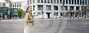 Vertical shot of young woman trying catch taxi, waving at driver on road, holding smartphone with car sharing app