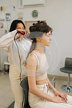 Vertical side shot of beautiful woman with black hair having it treated with curling iron by female hairdresser in