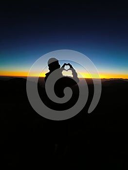 Vertical of a silhouette of a guy holding her hands in the shape of a heart against the sunset sky