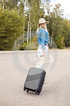 Vertical smiling blond woman hitchhiker with carton plate anywhere carrying roll aboard suitcase and waiting car stop photo