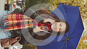 Vertical: Smiling woman with blue umbrella under rain talking on phone on street