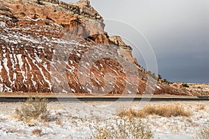 Vertical snow streaks on side of red rock mountain along empty highway