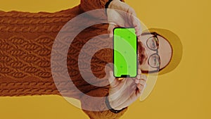 Vertical studio shot of a hipster millennial caucasian man showing his mockup green screen phone with two hands to the