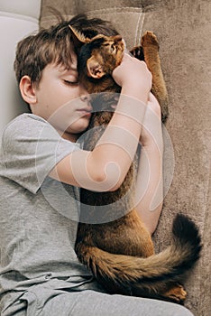 Vertical top view little kid boy lying on the couch and hugging his fluffy tail red cat kitten of somaly breed.