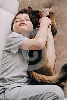 Vertical top view little kid boy lying on the couch and hugging his fluffy tail red cat kitten of somaly breed.