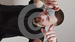 Vertical video - Attractive Caucasian man moves hands, microphone in hands, pointing to the camera grey background