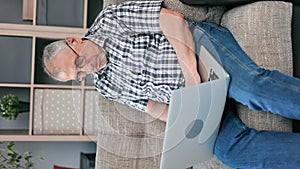 VERTICAL VIDEO POV focused elderly man working use laptop pc sitting on comfortable couch at home