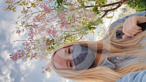 Vertical video. Voman wearing a facemask makes selfie video standing under blooming sakura tree. Travel to Japan in a