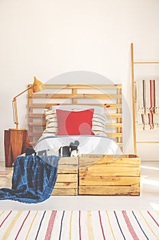 Vertical view of comfortable bed with wooden headboard, red pillow and bright bedding, bedroom interior with copy space on the whi