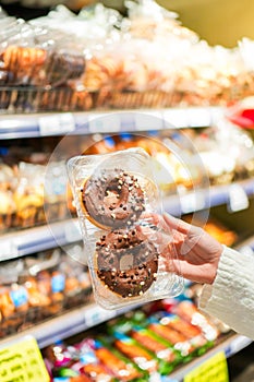 Vertical view of female hand showing to camera brown chocolate frosting with sprinkle doughnut in a supermarket while grocery